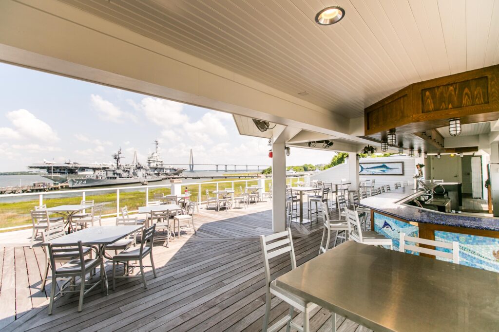 View of Charleston Harbor from The Bridge Bar at the Fish House. Looking toward The Ravenel Bridge and beyond the Yorktown Battleship. Tables and bar on an open air rooftop deck with ample seating and seaside decor.
