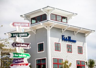 Image of the exterior of the Fish House. Gray siding with red trim in windows. Blue sign reading "Fish House" and directional signs out front out of focus.