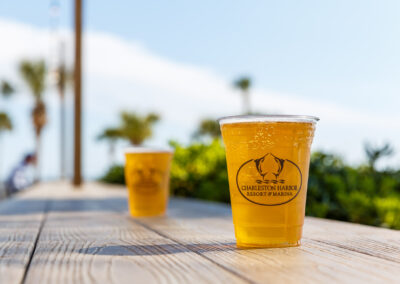 Two cold beers in Charleston Harbor Resort and Marina plastic cups sitting on a wooden bar top with palmetto trees out of focus in the background.